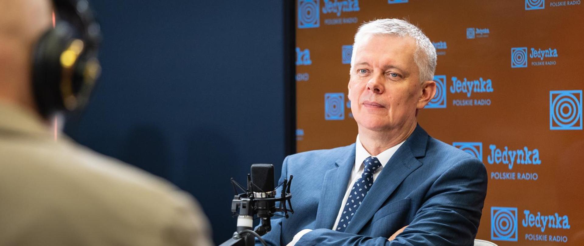 Minister Tomasz Siemoniak siedzi przed czarnym mikrofonem w studiu Programu 1 Polskiego Radia. W tle za ministrem - po prawej stronie pomarańczowa ścianka z logo i napisem Jedynka Polskie Radio (zmultiplikowane), a po lewej stronie granatowa ściana. Po lewej stronie zdjęcia widać siedzącego tyłem redaktora Grzegorza Osieckiego.