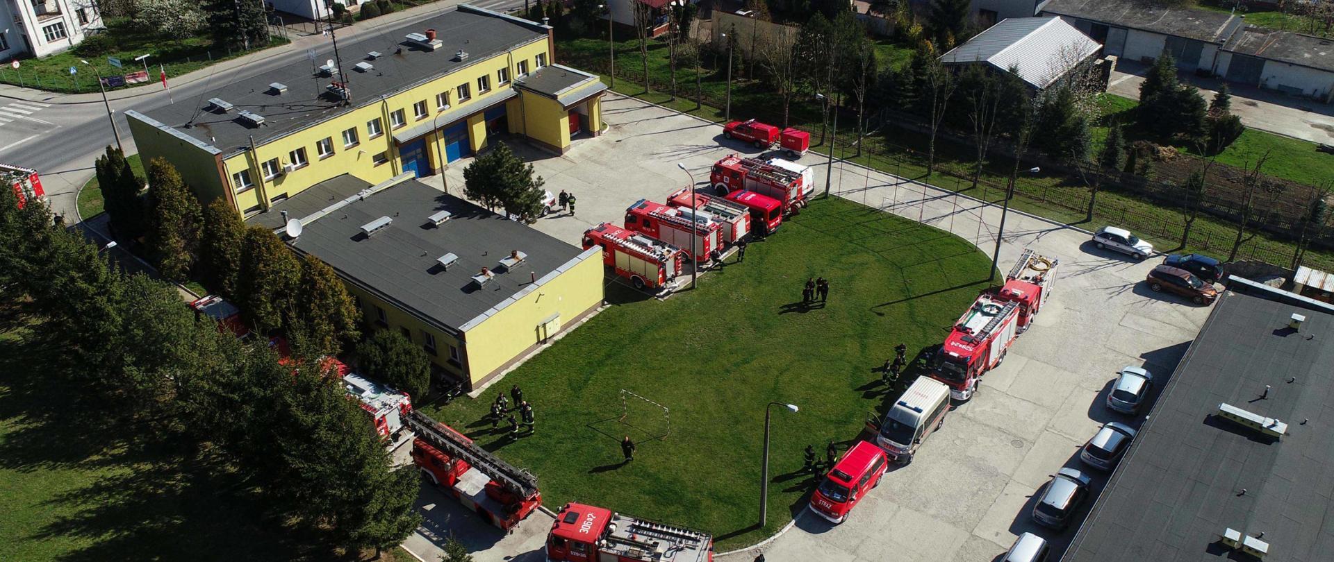 Zdjęcie zrobione wczesną wiosną w słoneczny dzień, dronem z lotu ptaka. Widoczne zabudowania komendy powiatowej i boisko trawiaste. Na placu wewnętrznym zaparkowane są samochody strażackie i osobowe. W tle widoczne sąsiednie zabudowania przy ul. 3 maja.