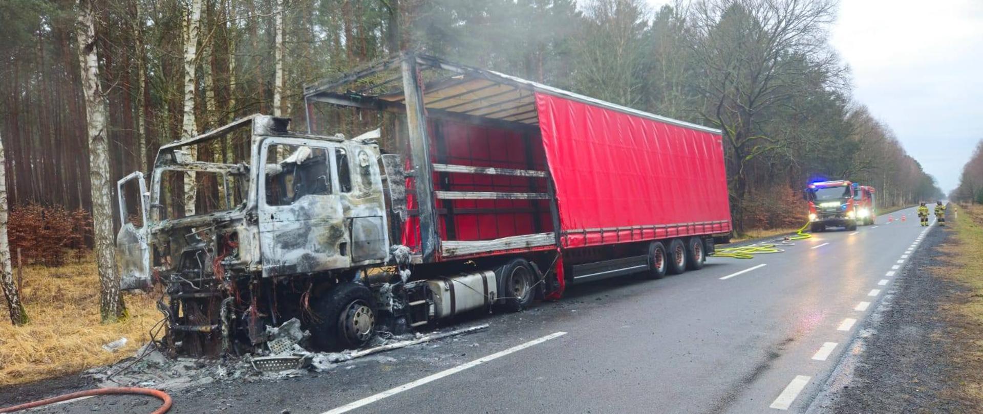 Samochód ciężarowy na drodze, spalony ciągnik siodłowy i część naczepy, w tle las i samochody pożarnicze
