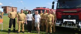 Grupa strażaków PSP oraz uczestników wydarzenia pozuje do wspólnego zdjęcia na boisku w Olszowie, na tle samochodów pożarniczych wykorzystywanych w działaniach ratowniczych.