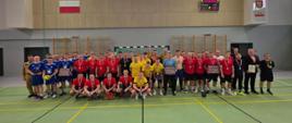 Uczestnicy finału Mistrzostw w Piłce Halowej Strażaków pozują do wspólnego zdjęcia na boisku w hali sportowej. Zawodnicy mają na sobie stroje sportowe w barwach swoich drużyn, a część z nich prezentuje medale i pamiątkowe dyplomy.