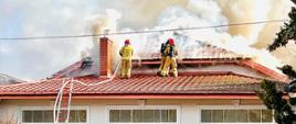 Na zdjęciu widać strażaków gaszących pożar budynku. Strażacy znajdują się na dachu budynku jak również przed nim. Z pod podbitki dachowej wydobywa się gęsty jasny dym. Strażacy ubrani są w odzież ochronną oraz aparaty ODO. Do budynku przystawiona jest drabina. W tle widać niebieski niebo.