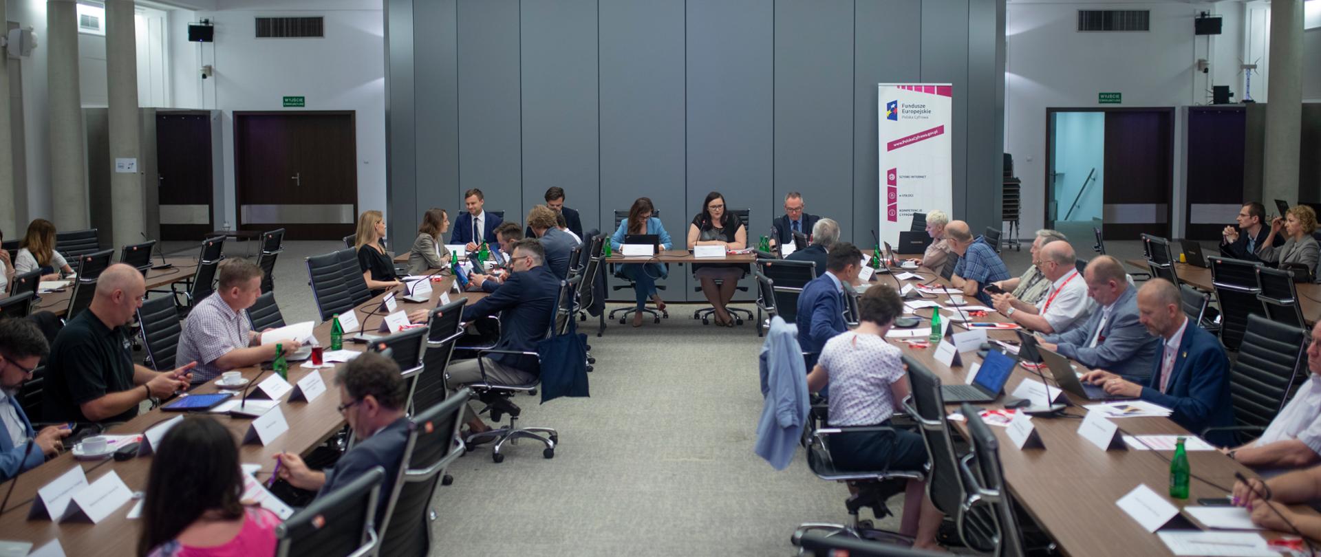Na zdjęciu sala konferencyjna, na środku ustawione dwa stoły, w poprzek na końcu sali stół prezydialny. Za stołem prezydialnym po prawej stronie roll-up z logo funduszy unijnych i nazwą Polska Cyfrowa.