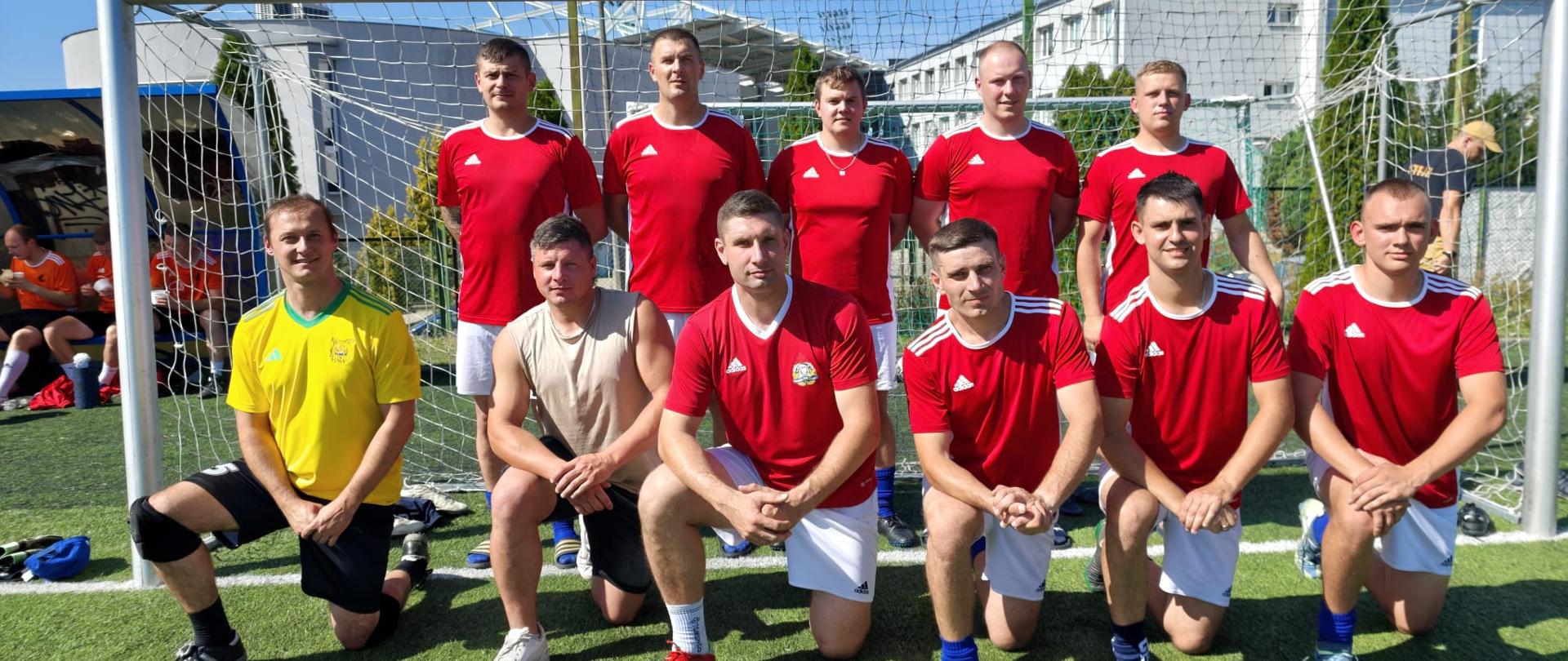 Wspólne zdjęcie funkcjonariuszy PSP - zawodników sportowych, którzy wzięli udział w Mistrzostwach w Piłce Nożnej. Zawodnicy ubrani są w biało czerwone stroje. Bramkarz ma stój w kolorze żółtym. Zdjęcie zrobione na tle brami na stadionie sportowym. 