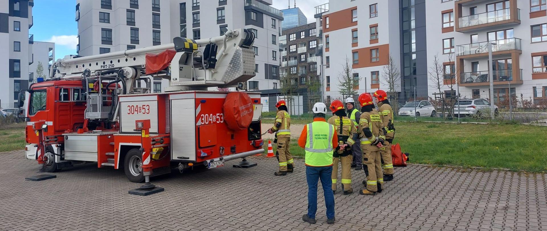 Strażacy w ubraniu typu nomex oraz przedstawiciele UDT uczestniczą w egzaminie przy podnośniku hydraulicznym