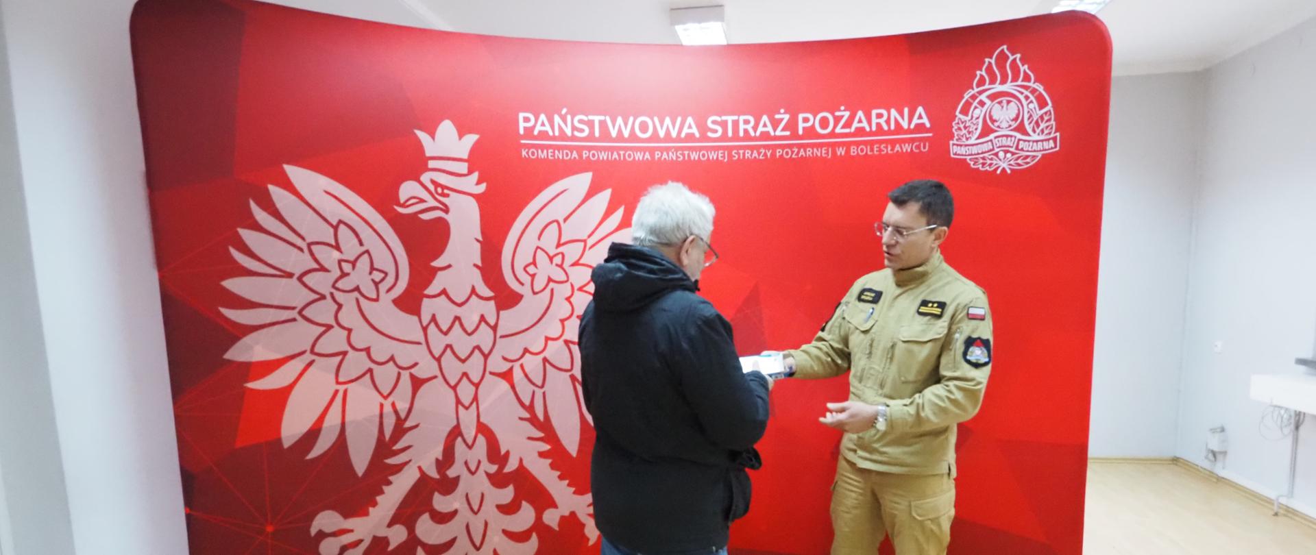 Zdjęcie przedstawia spotkanie dwóch mężczyzn na tle dużej, czerwonej ścianki reklamowej Komendy Powiatowej Państwowej Straży Pożarnej w Bolesławcu, na której widnieje godło Polski oraz logo straży. Po prawej stronie stoi strażak w piaskowym mundurze służbowym, który gestykuluje i rozmawia z mężczyzną stojącym przed nim tyłem do obiektywu, ubranym w ciemną kurtkę. Scena rozgrywa się w jasnym pomieszczeniu biurowym.