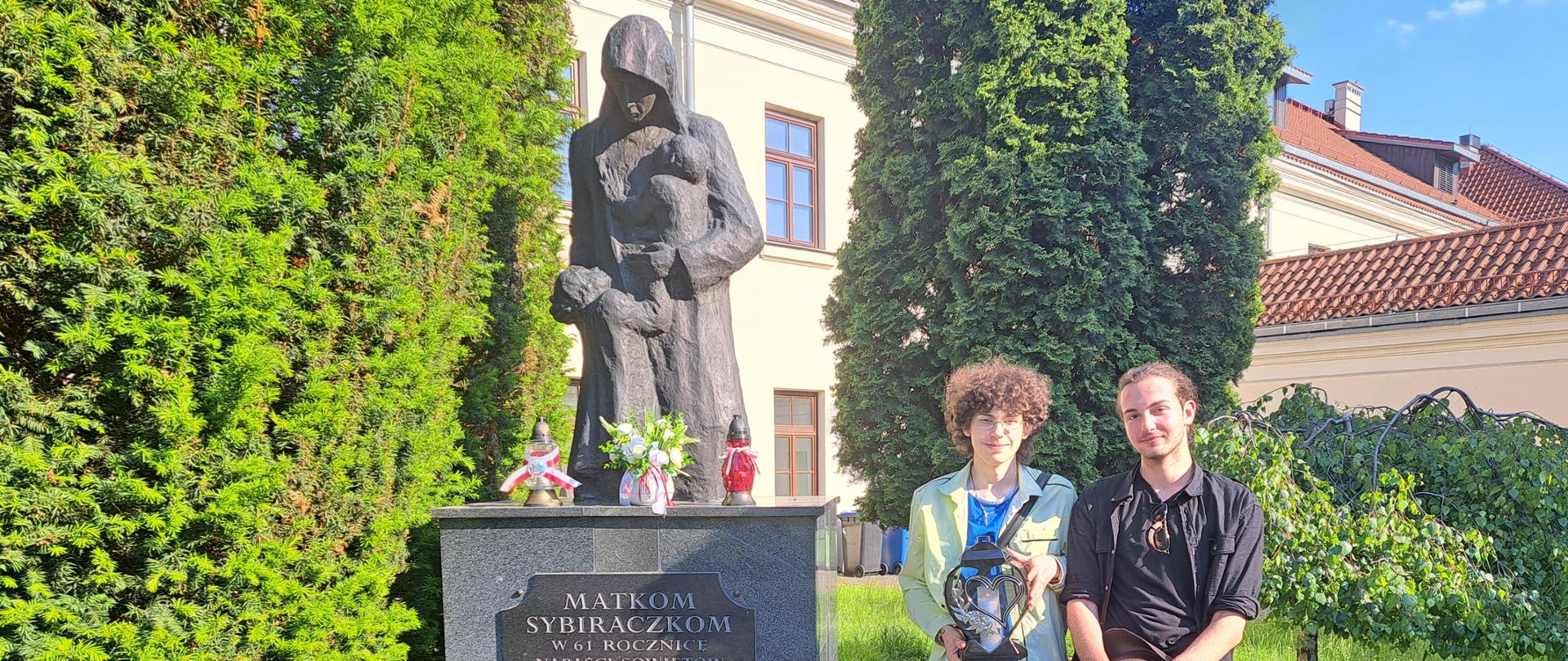 Z prawej strony pomnika "Matkom Sybiraczkom" stoi dwóch chłopców - jeden trzyma znicz, a drugi kapelusz. Pomnik jest udekorowany kwiatami białymi i czerwonymi, na cokole stoją znicze. w tle widać fragmenty budynku z czerwonym dachem, wokół zielone drzewa i kwiaty, z przodu wybrukowana ścieżka.