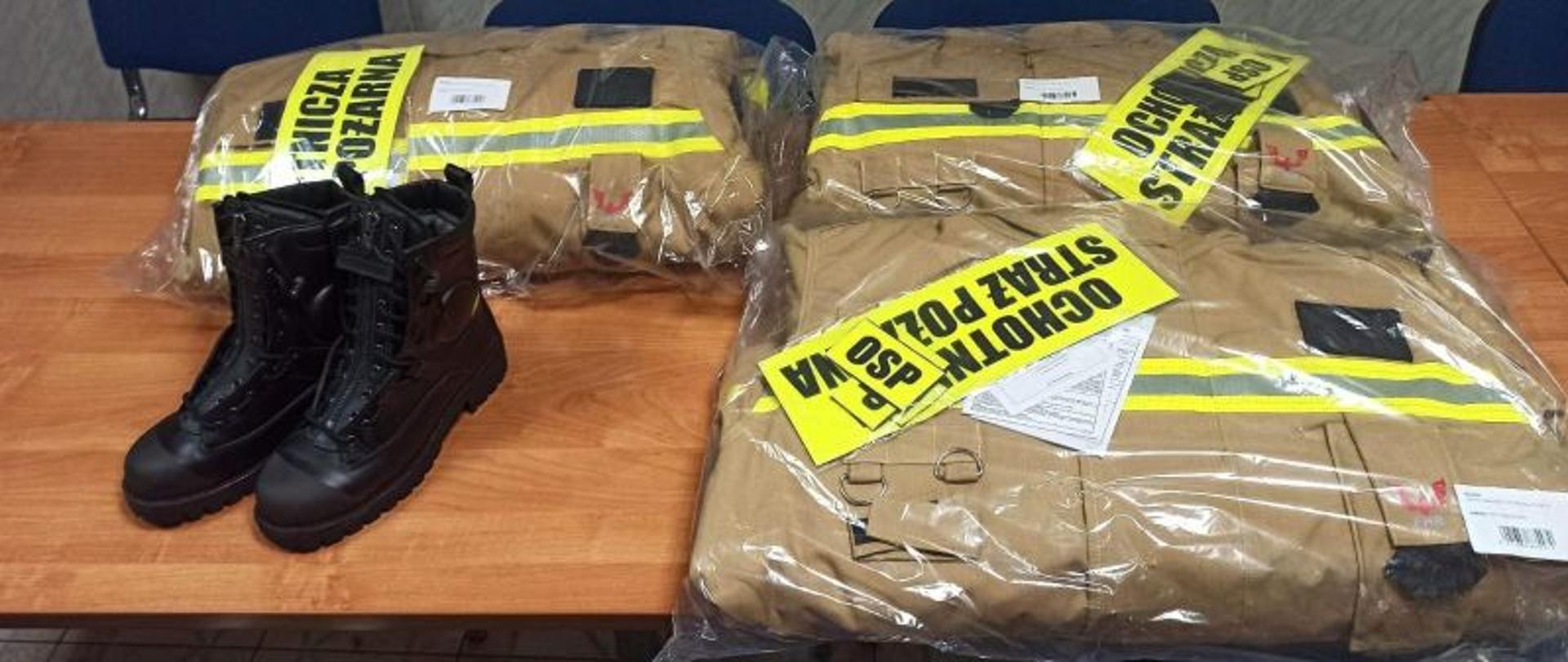 Zdjęcie przedstawia umundurowanie specjalne koloru piaskowego oraz buty specjalne. Sprzęt jest nowy, spakowany, lezy na stole.