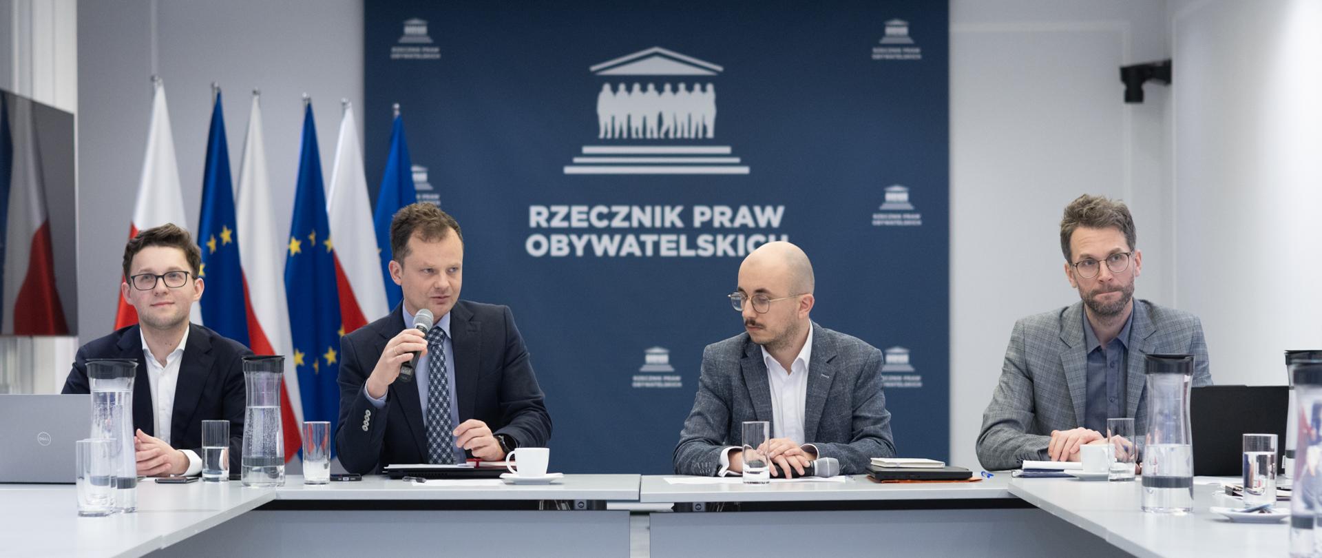 Cztery osoby siedzą w szczycie stołu konferencyjnego. Jedna z nich wypowiada się, trzymając w dłoni mikrofon. 