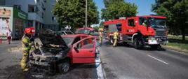 Strażak po lewej stronie zdjęcia dogasza prądem wody spaloną komorę silnika samochodu osobowego. Po prawej stronie zdjęcia widoczny samochód gaśniczy oraz dwóch strażaków.