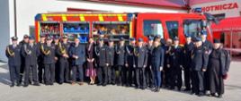 Zdjęcie grupowe wykonane na tle nowego pojazdu gaśniczego OSP Mścice. Na zdjęciu strażacy w galowych mundurach OSP i PSP. Księża, cywile. Wszyscy pozują do zdjęcia. Jest ładny dzień.