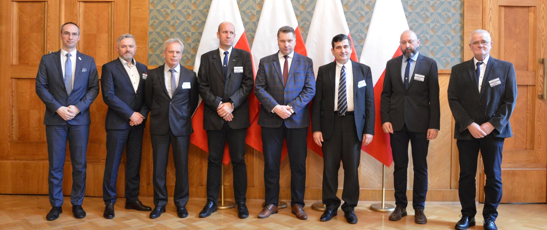 Ośmiu mężczyzn w garniturach, wśród nich minister Czarnek, stoi na tle zielonej ściany, za nimi cztery polskie flagi i drewniane drzwi.