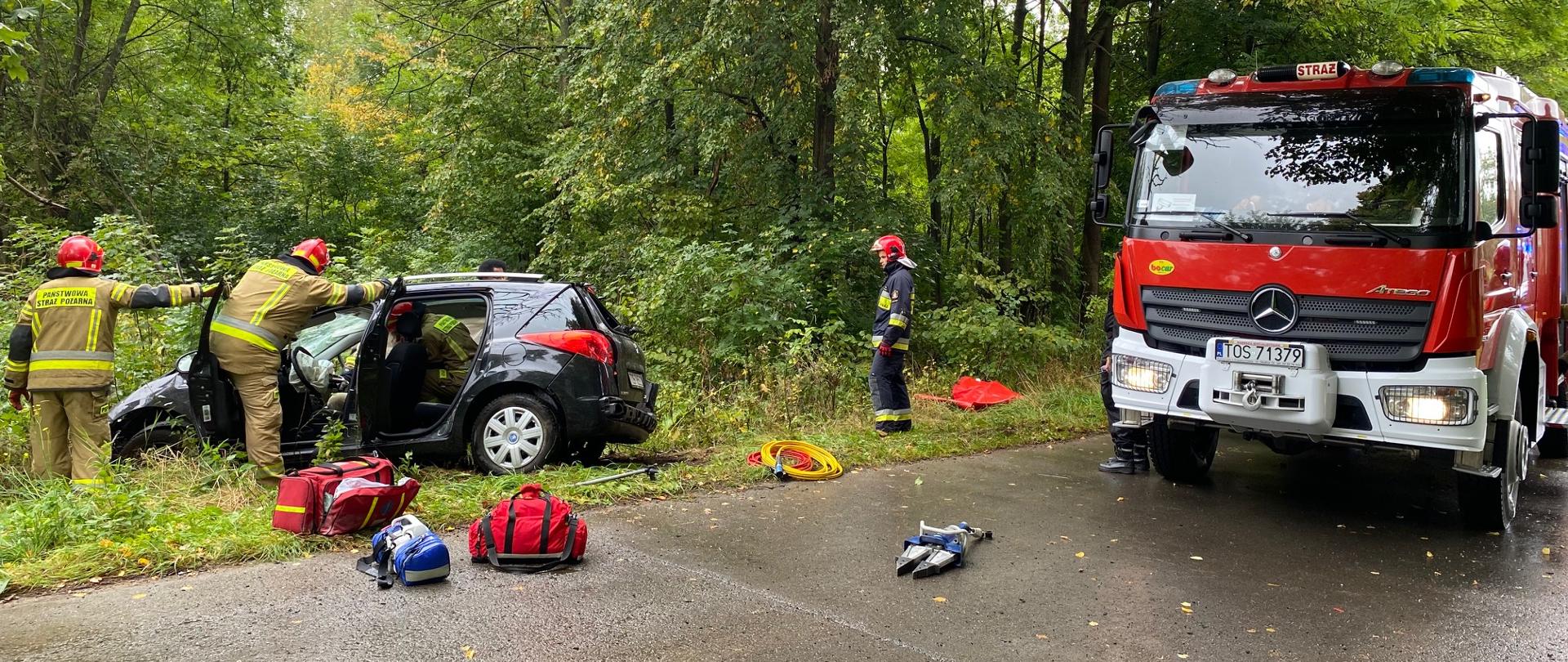 Na poboczu drogi stoi rozbity samochód Peugeot 207. Strażacy znajdujący się przy aucie zabezpieczają teren działania. Na jezdni zaparkowany samochód gaśniczy a przed nim leży sprzęt medyczny i norzyce hydrauliczne. 