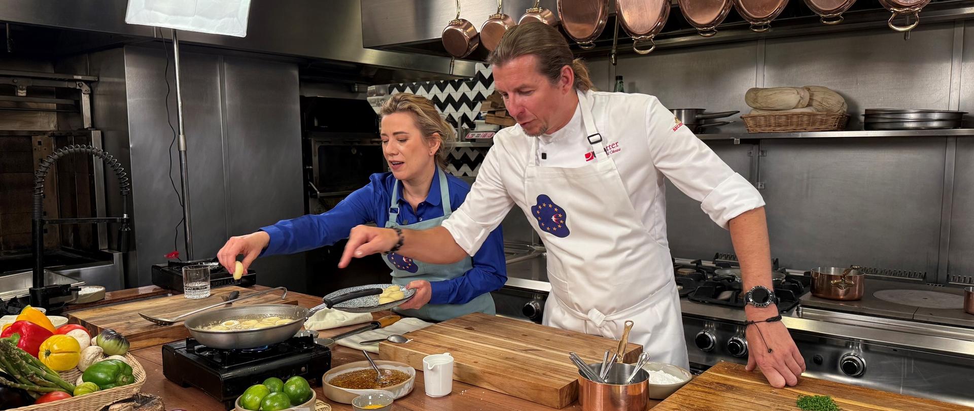 En la imagen se ve una cocina profesional de restaurante. Junto a una gran mesa de trabajo de madera están de pie dos personas vestidas con delantales de cocina, que cocinan juntas: la embajadora Agnieszka-Frydrychowicz-Tekieli y el chef Karol Okrasa.
En la sartén se fríen piezas redondas de comida en una salsa clara. Sobre la mesa, alrededor, hay muchos ingredientes: verduras de colores (pimientos, cebolla, champiñones), limas, hierbas, cuencos con salsas, ñoquis o pierogi crudos, cuchillos y tablas para cortar. Todo está cuidadosamente acomodado.