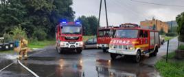 Zdjęcie przedstawia trzy samochody pożarnicze z jednostek Ochotniczych Straży Pożarnych z terenu gminy Wleń stojące na drodze. Na zdjęciu widoczny jest strażak oraz wąż pożarniczy, którym wypompowywana jest woda z budynku. 