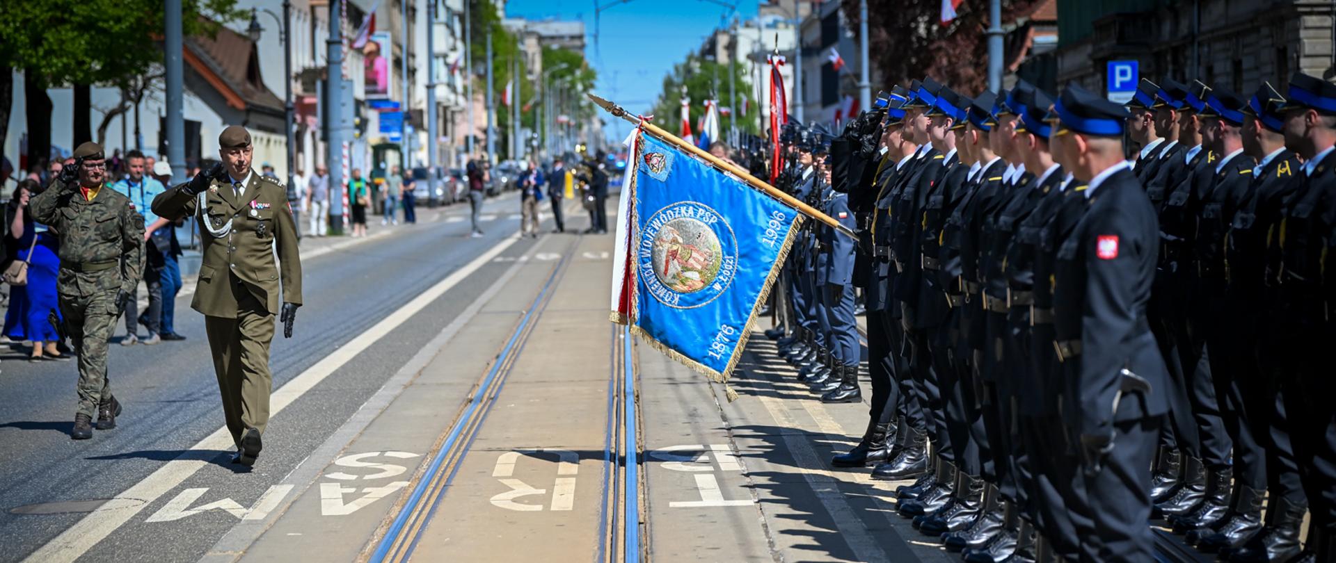 Oficerowie wojska dokonują przeglądu rzędu strażaków trzymających niebieski sztandar na ulicy z torowiskiem tramwajowym.