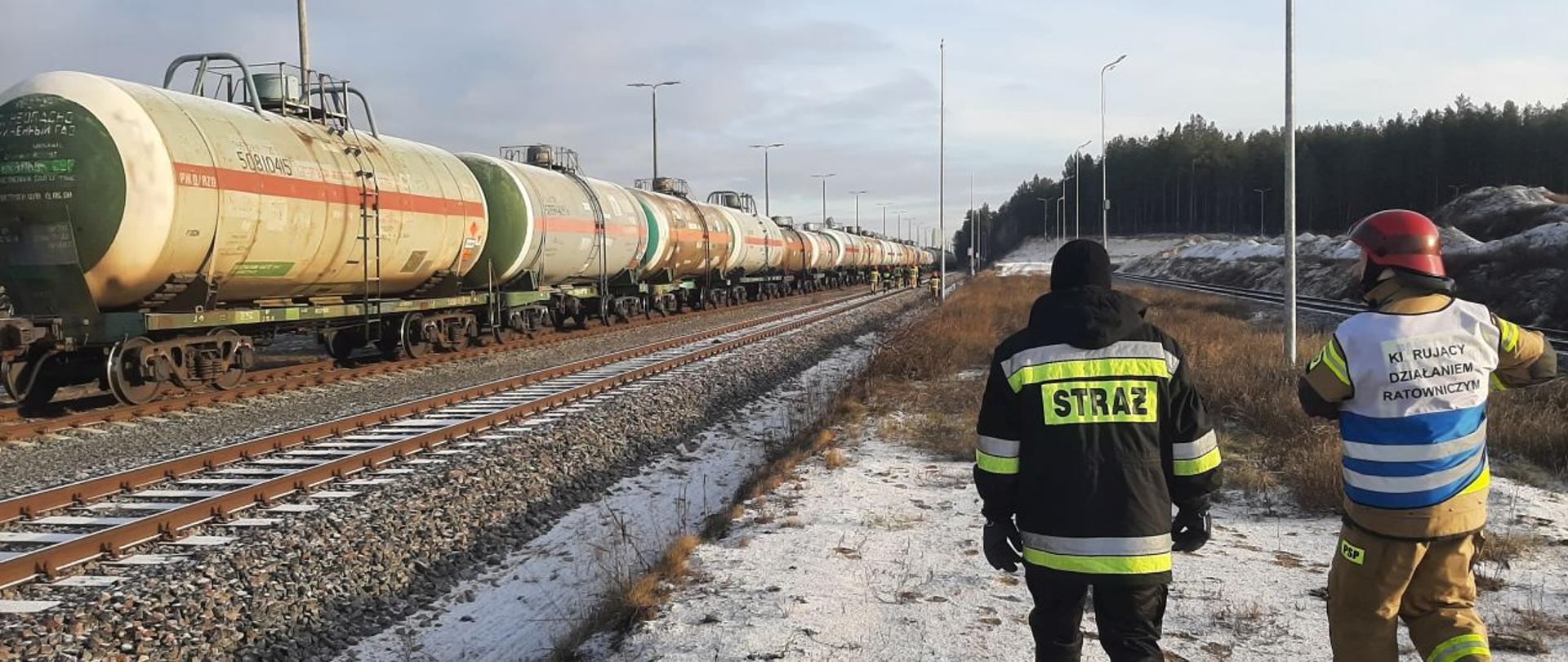 Widoczny skład kolejowy z cysternami, w tle strażacy pracujący przy uszkodzonym zaworze, widoczny dowódca akcji oraz oficer operacyjny