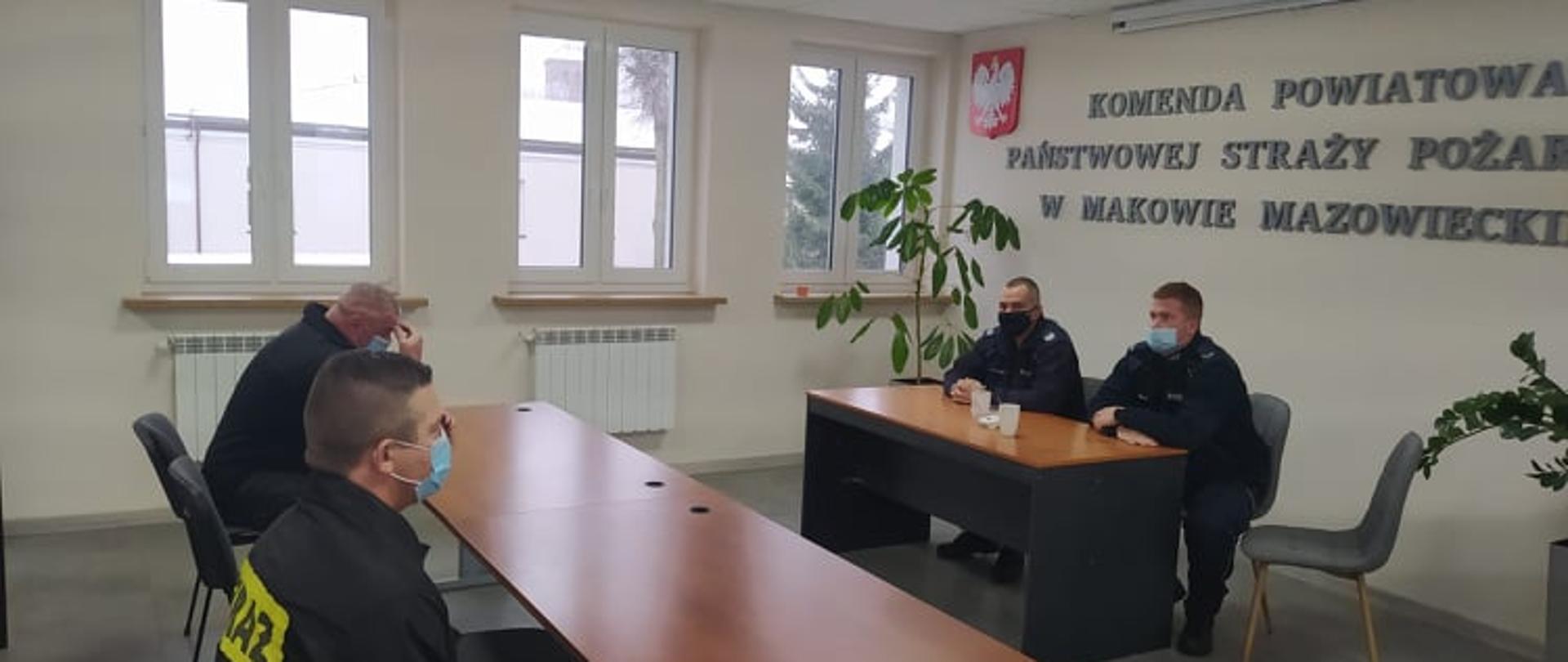 Dwóch funkcjonariuszy Policji siedzi przy stole na przeciwko strażaków zajmujących miejsca przy stolikach.