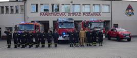 24 strażaków stoi w dwuszeregu przed pojazdami pożarniczymi, które mają włączone sygnały. Oddają honor.