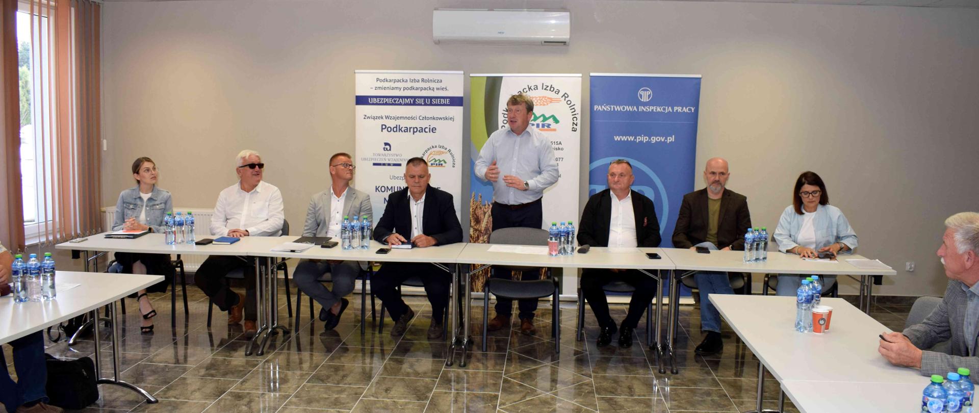 Przy jasnym stole konferencyjnym siedzi kila osób. Jeden mężczyzna stoi i przemawia. Na podłodze są brązowe płytki, ściana jest jasna. Za plecami siedzących ustawiono banery promocyjne Instytucji. 