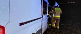 Kolizja na drodze wojewódzkiej. Zdjęcie przedstawia samochód typu bus, koloru białego, który uczestniczył w kolizji. Widoczne sa zniszczenia przedniej jego części. Obok samochodu stoi strażak w umundurowaniu koloru piaskowego i w białym hełmie. Strażak zagląda do wnętrza pojazdu. W oddali widoczne są świecące latarnie.