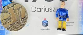 Zbliżenie na numer startowy z 17. PKO Poznań Półmaratonu z numerem „4736” i imieniem „Dariusz”. Obok numeru znajduje się medal z datą „13.04.2025” i napisem „PKO Poznań Półmaraton”, zawieszony na kolorowej wstążce w odcieniach różu, fioletu i niebieskiego. Po prawej stronie leży mała figurka mężczyzny w niebieskiej koszuli i żółtych spodniach, przypominająca zabawkowego strażaka lub ratownika. Tło stanowi fragment wzoru z grafikami budynków, nawiązujący do miejskiej tematyki biegu.