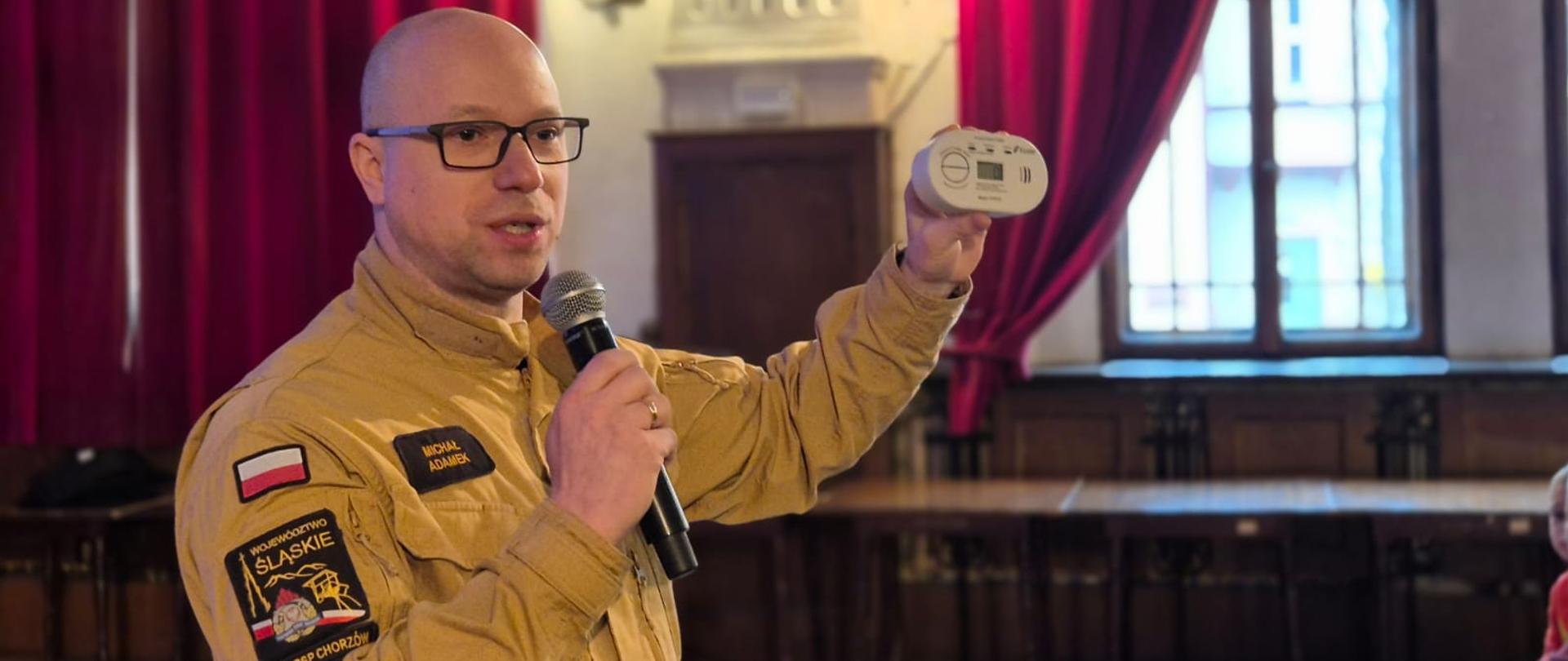 Na zdjęciu widać strażaka w beżowym mundurze, stojącego w sali z czerwonymi zasłonami i dużymi oknami. Mężczyzna trzyma mikrofon oraz czujnik dymu lub tlenku węgla, prezentując go uczestnikom spotkania. Na mundurze widoczne są naszywki, w tym flaga Polski oraz oznaczenie Komendy Miejskiej PSP w Chorzowie. Strażak prowadzi prelekcję w ramach działań edukacyjnych dotyczących bezpiecznych ferii.