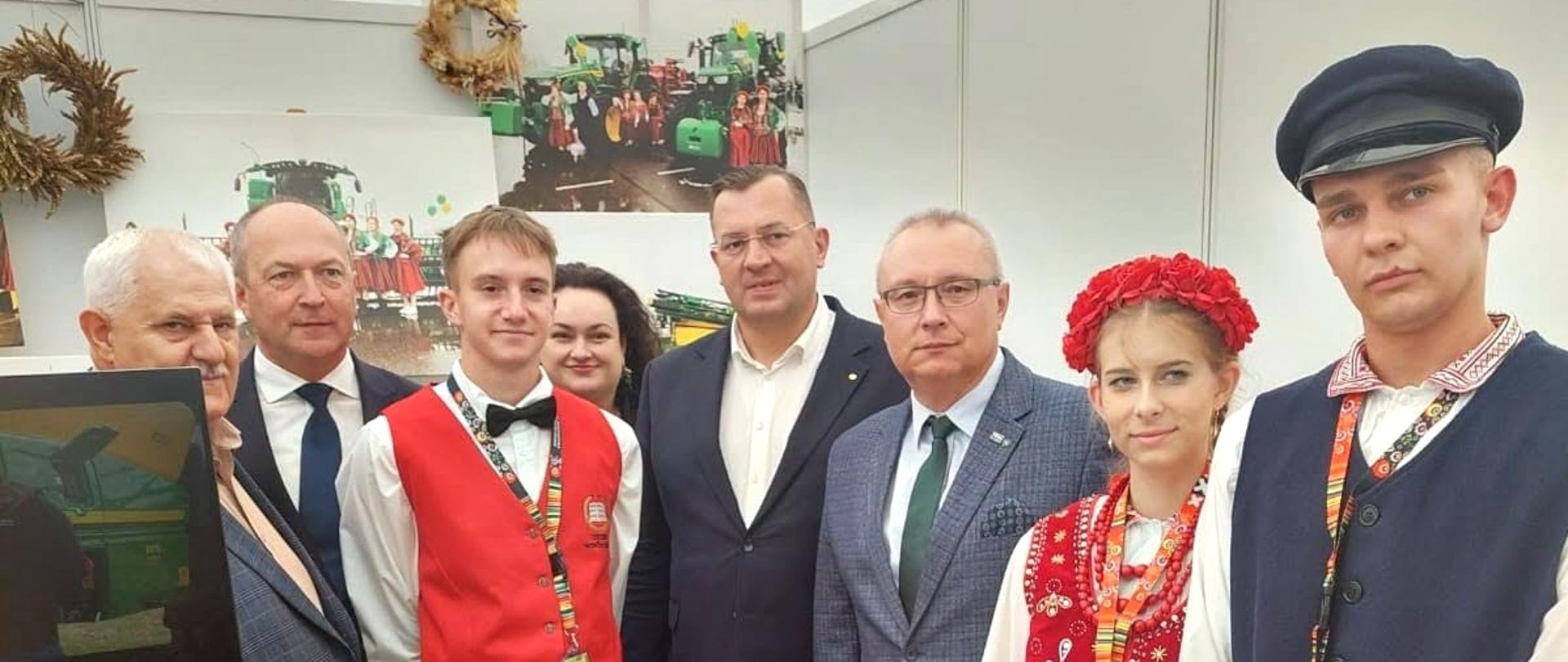 Grupa uczniów i dorosłych uczestników podczas XXV Krajowej Wystawy Rolniczej w Częstochowie, na stoisku Branżowego Centrum Umiejętności. W centrum stoją przedstawiciele ministerstwa, a po bokach dwoje uczniów w strojach ludowych. W tle znajdują się zdjęcia związane z rolnictwem i tradycjami polskiej wsi.