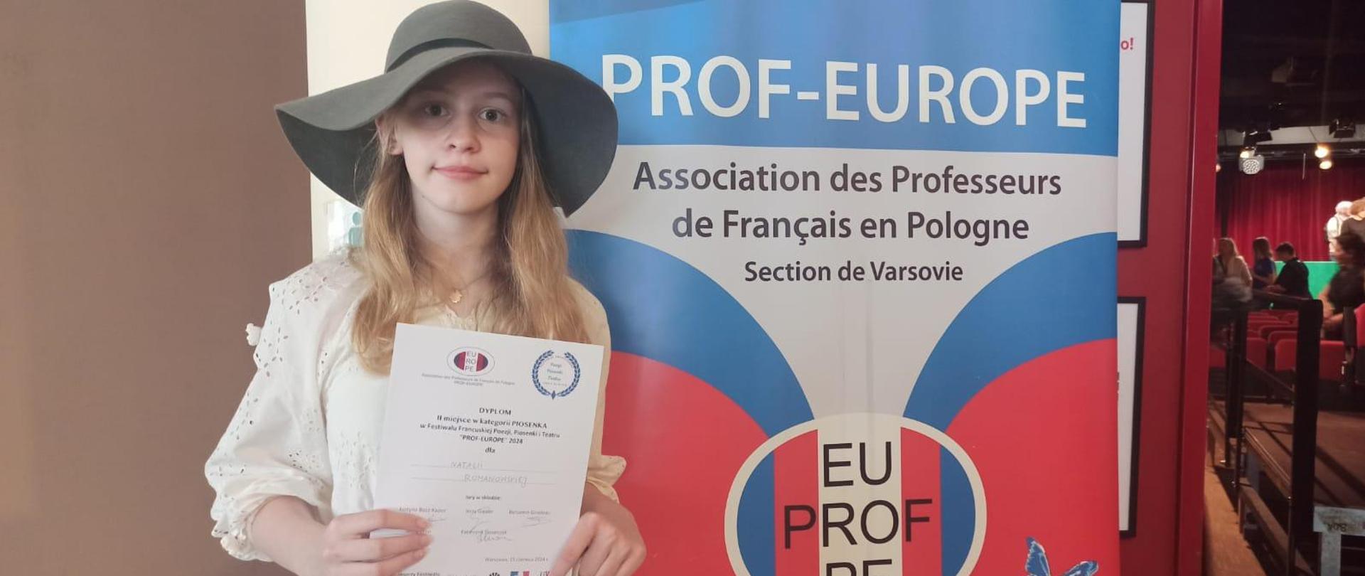 Dziewczynka ubrana w białą sukienkę z ciemnym kapeluszem na głowie, w rękach trzyma dyplom. Dziewczynka stoi na tle kolorowego baneru z nazwą konkursu.
