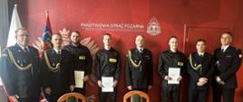 Pamiątkowe zdjęcie grupowe nowo zaprzysiężonych strażaków z kierownictwem KM PSP Szczecin na tle ścianki z logotypem Państwowej Straży Pożarnej. Strażacy trzymają akty ślubowania.