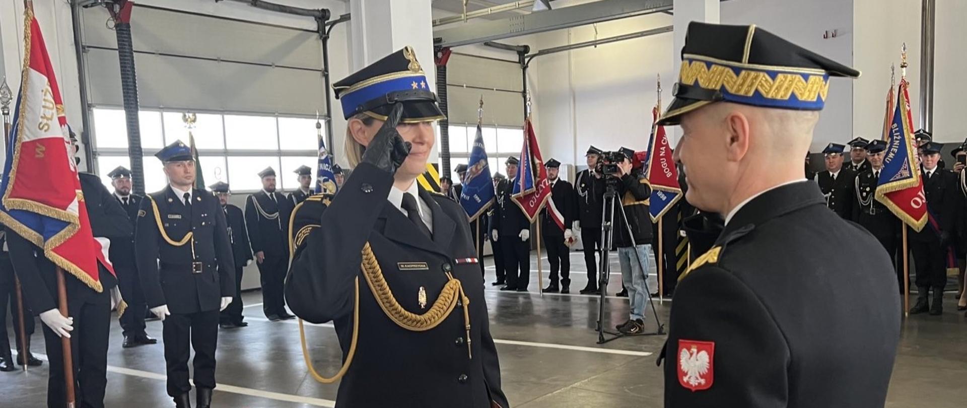 Uczestnicy uroczystości stojący w garażu jednostki ratowniczo - gaśniczej w Gnieźnie. Na pierwszym planie Pani komendant stojąca na czerwony dywaniarze w trakcie oddawania honoru komendantowi głównemu PSP. W tle poczty sztandarowe. 