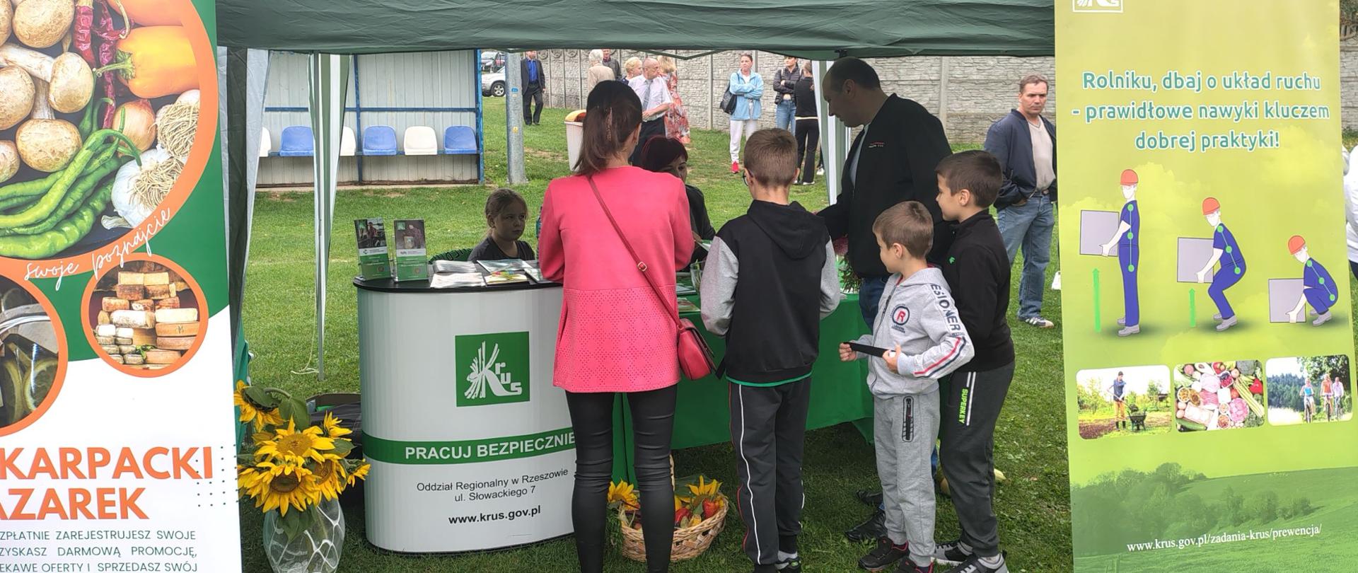 Przy stoisku informacyjno-promocyjnym Kasy znajduje się grupa osób – dwoje dorosłych i trójka dzieci. Na stoliku przed stoiskiem znajdują się materiały prewencyjne Kasy, a pracownik KRUS udziela informacji osobom odwiedzającym stoisko.