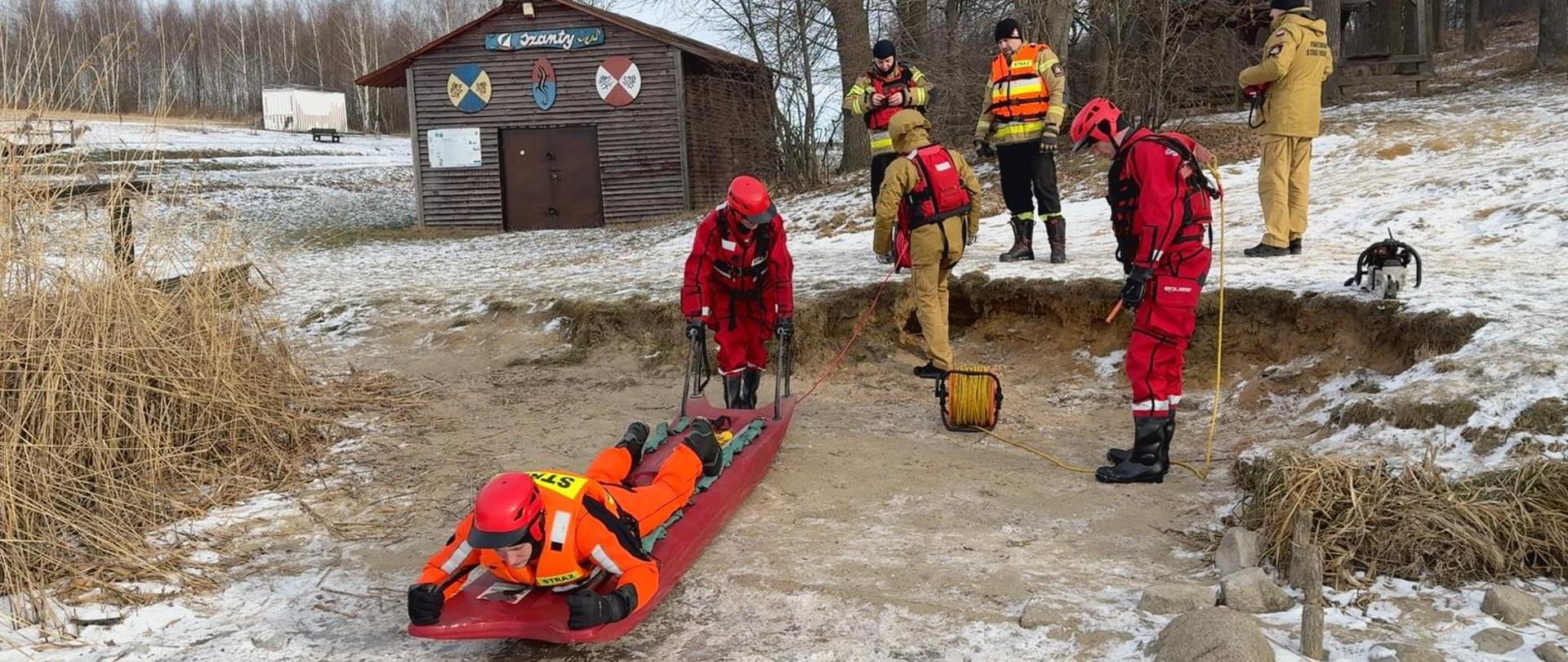 Na zdjęciu grupa ratowników w czerwonych i żółtych kombinezonach prowadzi zimowe ćwiczenia ratownicze. Kilku z nich stoi na zaśnieżonym terenie, a inni obsługują czerwone sanie, na których leży osoba udająca poszkodowanego. Sanie są ciągnięte za pomocą lin. W tle widać drewniany budynek oraz bezlistne drzewa, co podkreśla chłodne, zimowe warunki