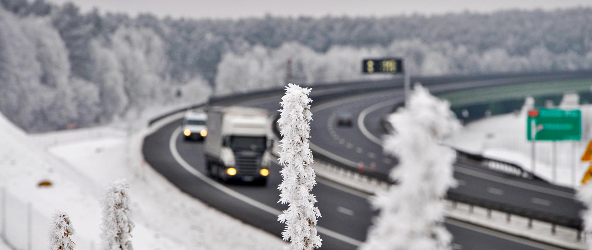 Zdjęcie przedstawia zakręt na drodze ekspresowej S3. Nawierzchnia drogi jest odśnieżona i czarna. Po drodze poruszają się różne pojazdy. Cały teren poza drogą pokryty jest warstwą śniegu. Na pierwszym planie roślina pokryta gęsto szronem.