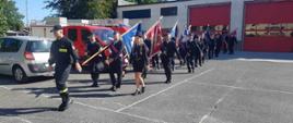 Szkolenie pocztów sztandarowych. Zdjęcie przedstawia plac komendy w Rawiczu. Na placu maszerują jeden za drugim poczty sztandarowe OSP. Na ich czele strażak-dowódca z PSP. Z lewej strony zaparkowane są auta. W tle budynek i drzewa.
