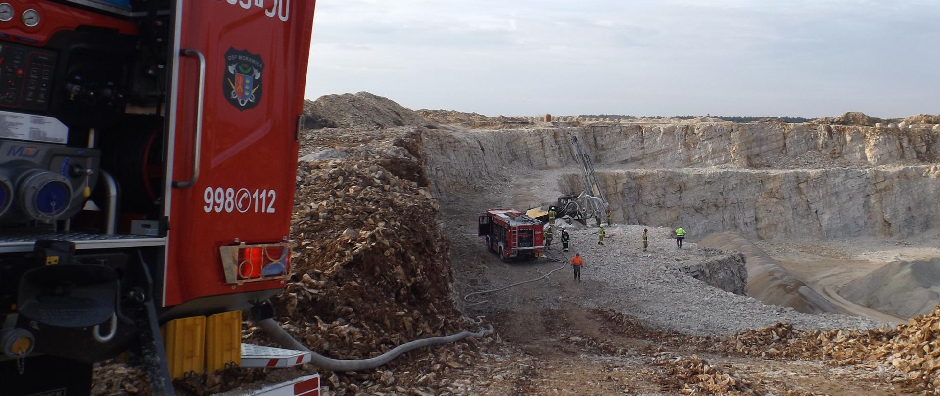 Na zdjęciu przedstawiono kopalnię wapienia gdzie miał miejsce pożar maszyny wiertniczej. Na pierwszym planie jest fragment samochodu pożarniczego biorącego udział w zdarzeniu, w tle wyrobisko kopalni, a w głębi widać drugi samochód gaśniczy, zniszczoną wiertnię i grupę strażaków.