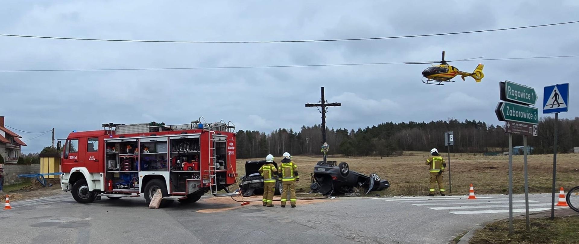 Zdjęcie przedstawia miejsce wypadku: z lewej strony stoi wóz strażacki, w centrum widać oba zniszczone samochody osobowe, a na ich tle dwóch ratowników. Z lewej strony startuje śmigłowiec Lotniczego Pogotowia Ratunkowego.
