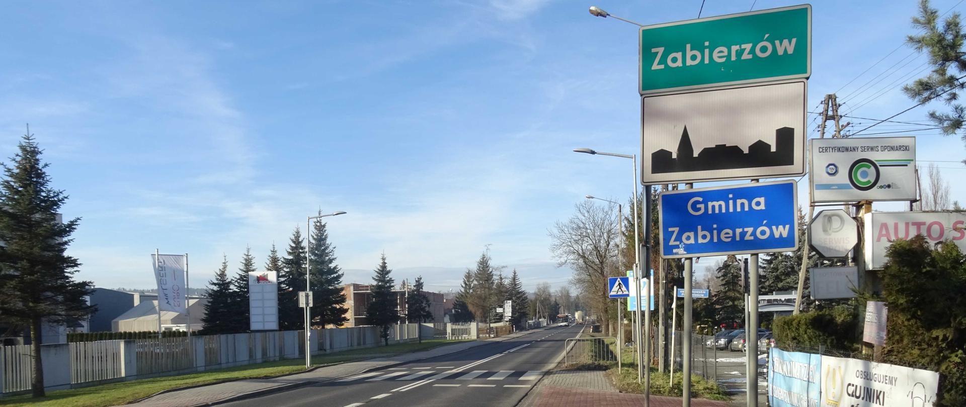 DK79 w Zabierzowie, po obu stronach chodnik i zabudowania, w oddali autobus i ciężarówka