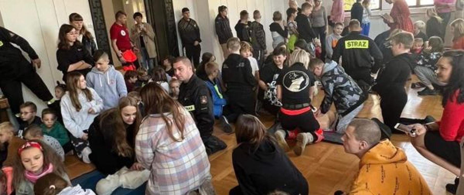 Widok na dużą salę. Sala jest pełna dzieci i dorosłych. Wszyscy skupieni na jednoczesnym wykonywaniu RKO.Wśród ludzi widoczni strażacy w czarnych mundurach. W tle widać białe ściany sali i duże okno.