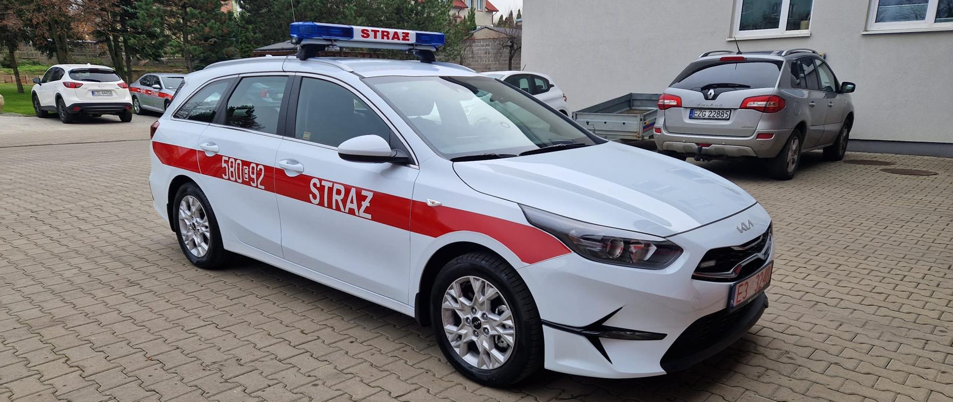 Zdjęcie przedstawia biały samochód z czerwoną taśmą na boku. powyższy samochód stoi na kotce brukowej na tle budynku komendy powiatowej PSP w Zgierzu.