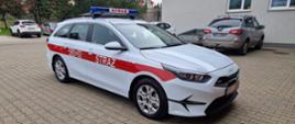 Zdjęcie przedstawia biały samochód z czerwoną taśmą na boku. powyższy samochód stoi na kotce brukowej na tle budynku komendy powiatowej PSP w Zgierzu.
