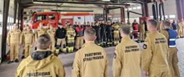 Umundurowani strażacy biorą udział w odprawie. Stoją na baczność, w tle stoi wóz strażacki.