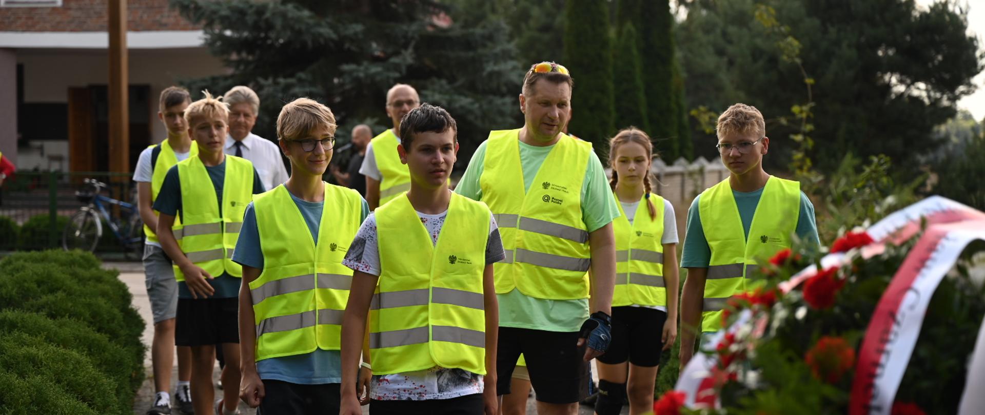 Po drodze wyłożonej szarą kostką idzie grupa osób w żółtych odblaskowych kamizelkach, ludzie z przodu niosą wieniec biało-czerwonych kwiatów.