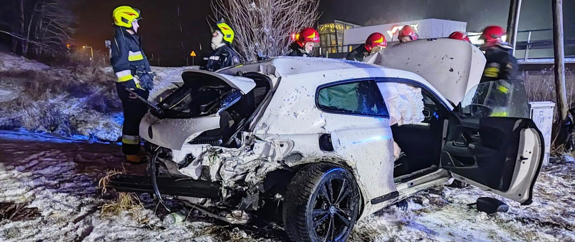 Uszkodzony samochód osobowy poza drogą a obok niego strażacy 