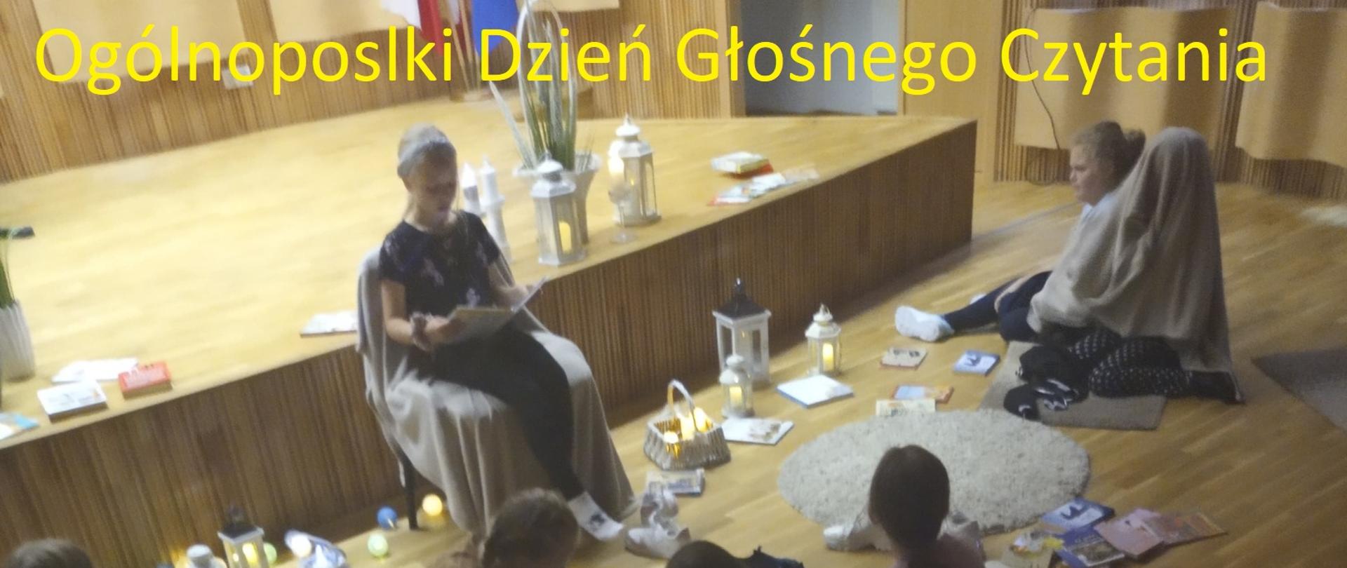 Zdjęcie przedstawia czytające dziecko w sali koncertowej. Wokół siedzą inne dzieci słuchające czytanej książki.