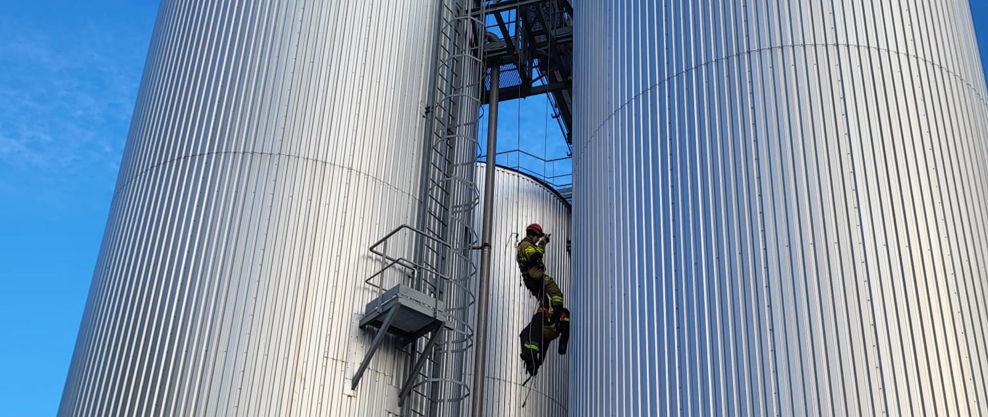 Strażacy przy pomocy lin opuszczają osobę poszkodowaną z metalowych silosów.