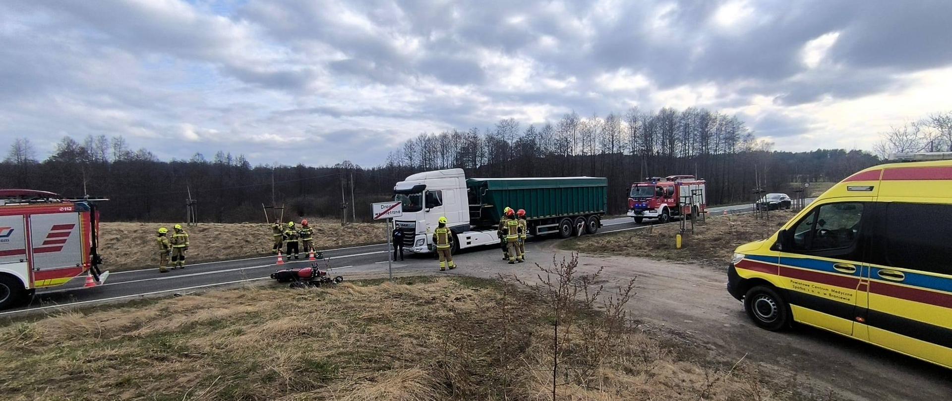 Na zdjęciu miejsce zdarzenia drogowego. Z prawej strony stoi żółta karetka pogotowia, na środku zdjęcia duża ciężarówka z naczepą i z przodu i z tyłu niej samochody strażackie. Strażacy przy samochodach.