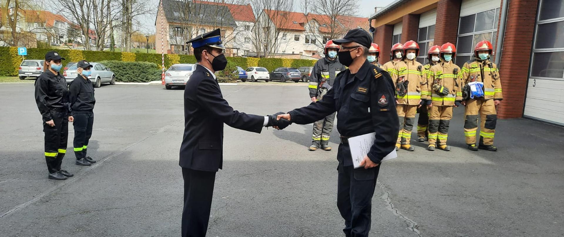 Na zdjęciu Komendant ubrany w mundur dowódczo-sztabowy wręcza strażakowi powołanie. Strażak ubrany w mundur wyjściowy. W tle po lewej stronie dwie funkcjonariuszki straży ubrane w mundur koszarowy, po prawej strażacy podziału bojowego ubrani w ubrania specjalne oraz czerwone hełmy.