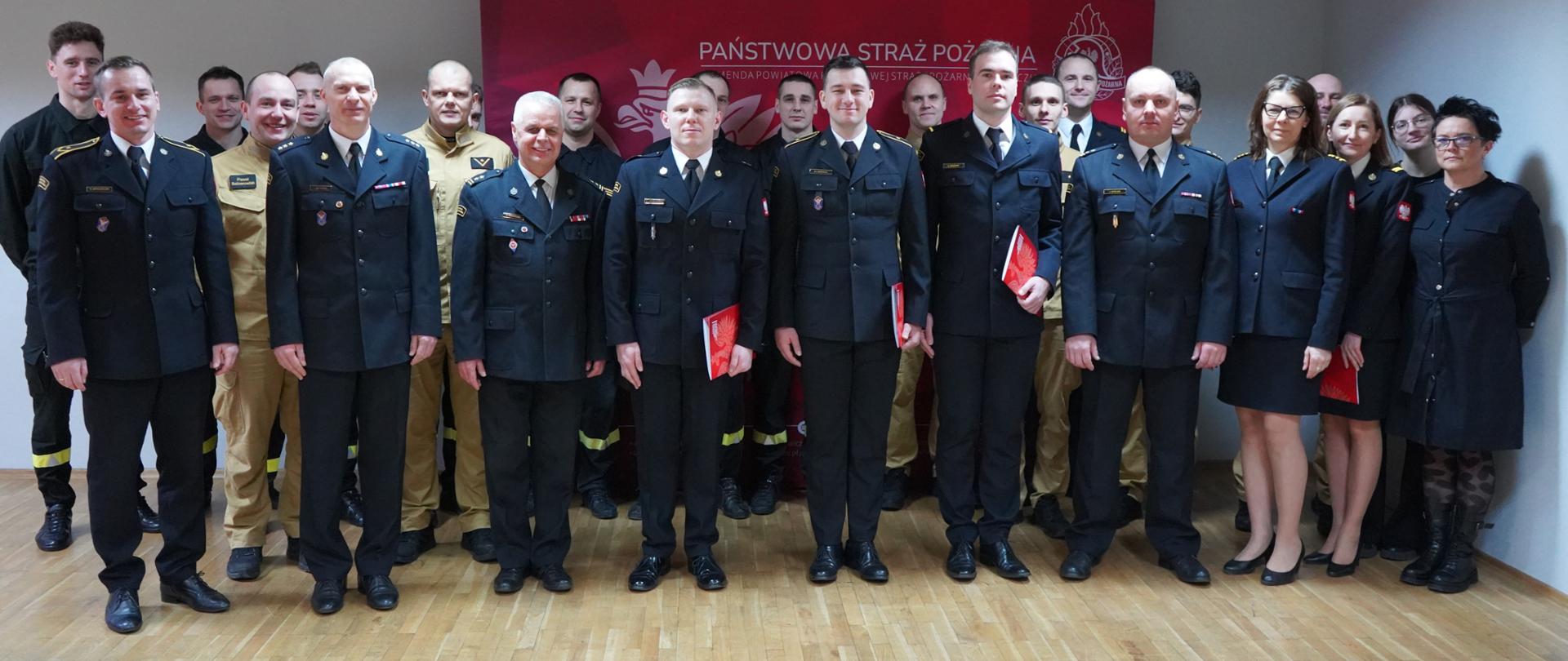 Świetlica Komendy Powiatowej PSP w Rawiczu. Na tle ścianki promocyjnej do wspólnego zdjęcia pozują wszyscy uczestnicy uroczystości. Wsród nich są zarówno strażacy jak i pracownicy cywilni. Część strażaków ma na sobie mundury wyjściwe, część koszarowe w kolorze ciemnym, inni - mundury służbowe w kolorze piaskowym. 