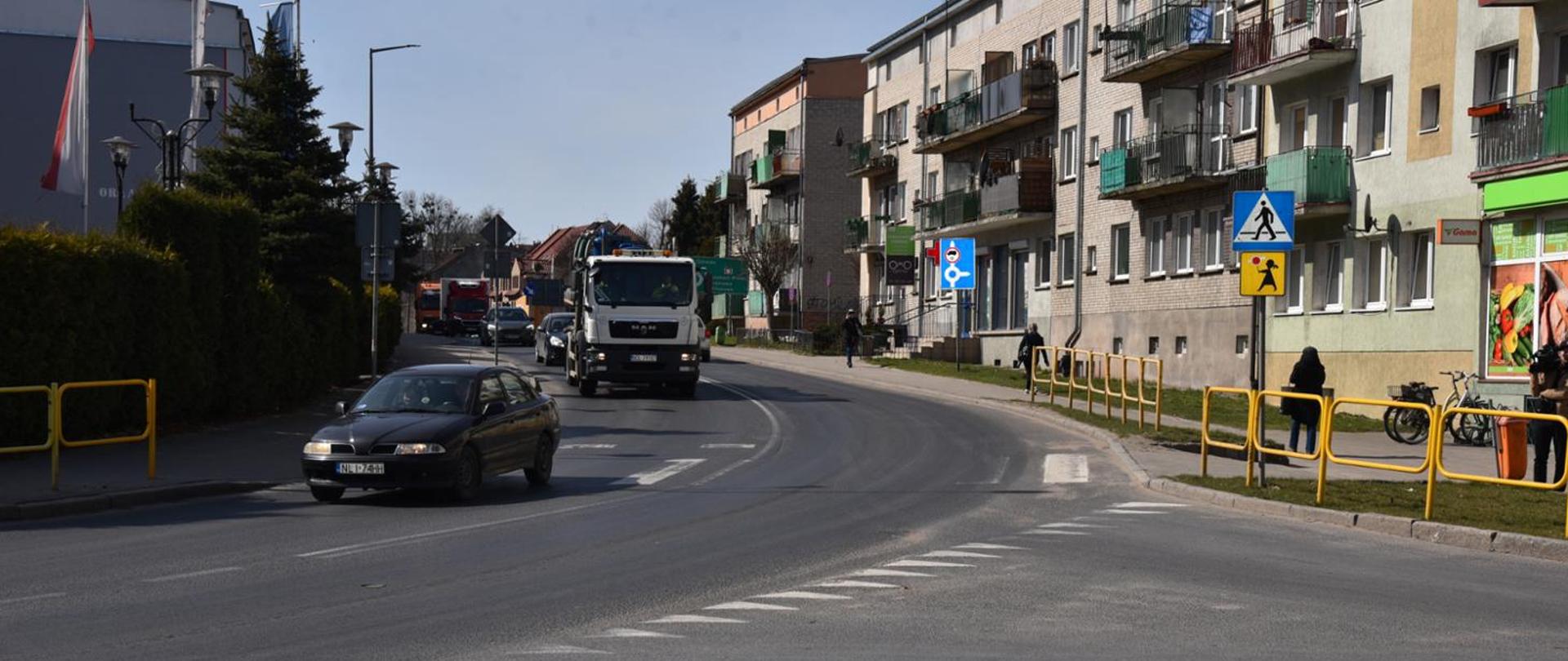 Bezchmurny dzień. Zakręt w mieście, którym jadą samochody osobowe i ciężarowe. Pojazd na pierwszym planie przejeżdża przez przejście dla pieszych.